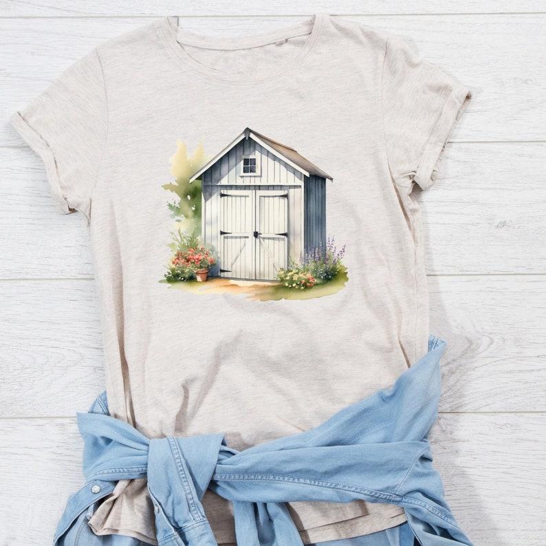 May include: A beige t-shirt with a watercolor illustration of a white shed with a door and a window. The shed is surrounded by green plants and flowers.