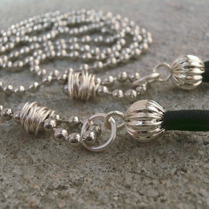 May include: A silver chain necklace with a black cord and two silver charms. The charms are round and have a textured surface.