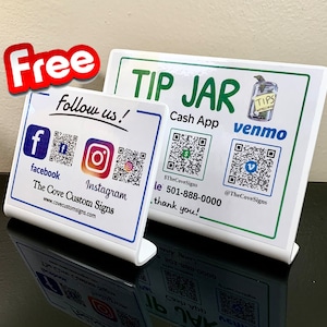 Könnte beinhalten: Ein weißes Schild mit grünem Text, der "TIP JAR" lautet und ein Cartoon-Bild eines Glases mit dem Wort "TIPS" darauf enthält. Das Schild enthält auch QR-Codes für Cash App und Venmo sowie den Text "$TheCoveSigns" und "@TheCoveSigns". Ein zweites weißes Schild mit blauem Text lautet "Follow us!" und enthält Social-Media-Symbole für Facebook und Instagram sowie den Text "The Cove Custom Signs" und "www.covecustomsigns.com".