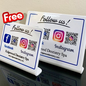 May include: Two white signs with blue borders, each with a QR code and the text "Follow us!" One sign has a Facebook logo and the text "Ridgewood Dentistry Spa" and the other has an Instagram logo and the text "Ridgewood Dentistry Spa".
