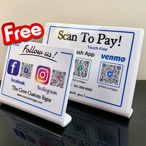 May include: Two white table top signs with blue borders. The top sign says "Scan To Pay!" with the text "Touch Free" and has QR codes for Cash App and Venmo. The bottom sign says "Follow us!" with QR codes for Facebook and Instagram. The text "The Cove Custom Signs" and a website address are also on the sign.