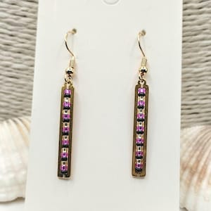 May include: A pair of gold-colored dangle earrings. Each earring features a rectangular, bronze-colored bar adorned with a vertical row of small, pink and black beads. The earrings hang from gold-colored fish hook ear wires.