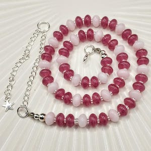 May include: A necklace with alternating pink and burgundy beads. The beads are strung on a silver-coloured chain with a clasp. A silver-coloured chain with a star charm extends from the necklace.