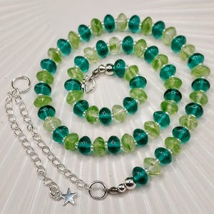 May include: A necklace with green and clear glass beads, strung together with a silver-tone chain. The beads alternate in colour, creating a striking contrast. The necklace has a star charm and a lobster clasp. The beads are a mix of light and dark green.