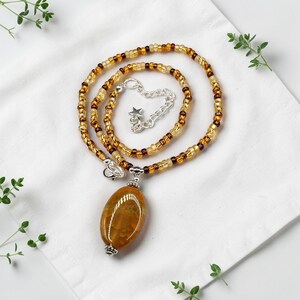 May include: A handmade necklace with an amber-coloured, oval pendant. The beaded chain is gold and brown, with a silver-toned clasp and a small star charm. The necklace is displayed on a white surface.