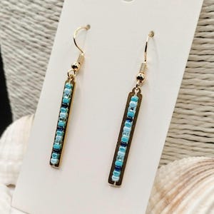May include: A pair of gold-coloured dangle earrings. Each earring has a rectangular gold-toned bar with a vertical row of small, colourful beads in shades of blue and turquoise. The earrings hang from a simple gold hook.