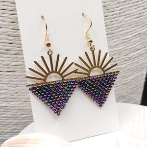 May include: A pair of gold-coloured earrings featuring a sunburst design above a triangle of iridescent beads. The earrings have a hook closure and are displayed on a white card.