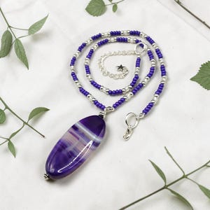 May include: A necklace featuring a large, oval purple and white agate pendant. The pendant hangs from a double strand necklace with silver and purple beads. The necklace has a silver clasp and a star charm.