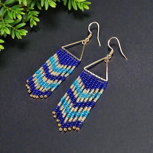 Peut inclure: Une paire de boucles d'oreilles en perles avec un motif géométrique. Les boucles d'oreilles présentent un dessus triangulaire doré avec une frange de perles bleues, bleu clair et dorées. Les boucles d'oreilles sont présentées sur une surface sombre.