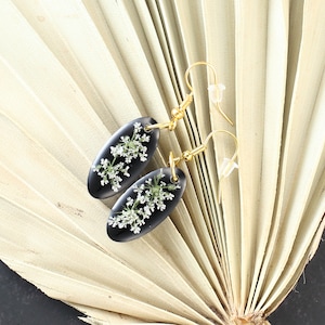 May include: A pair of black oval earrings with gold hooks. Each earring features a delicate pressed flower design with white petals.