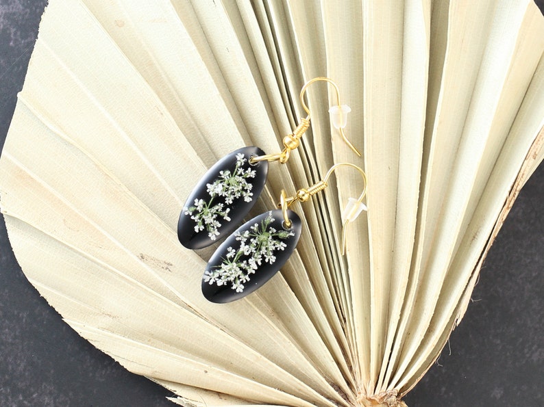May include: A pair of black oval earrings with gold hooks. Each earring features a delicate pressed flower design with white petals.