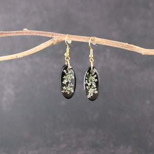 May include: A pair of black oval earrings with gold hooks. Each earring features a delicate white flower pressed inside the resin.
