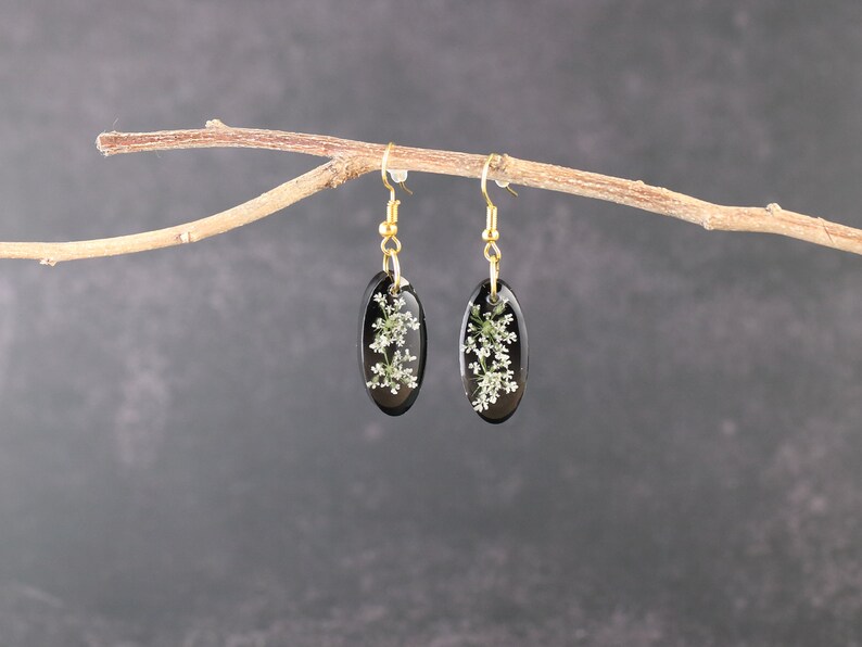 May include: A pair of black oval earrings with gold hooks. Each earring features a delicate white flower pressed inside the resin.