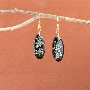 May include: A pair of black oval earrings with gold hooks. Each earring features a delicate white flower pressed inside the resin.