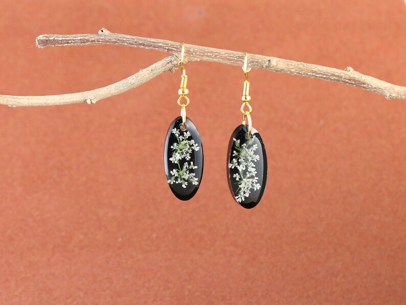 May include: A pair of black oval earrings with gold hooks. Each earring features a delicate white flower pressed inside the resin.