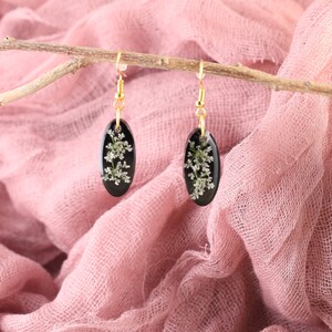 May include: A pair of black oval earrings with gold hooks. Each earring features a delicate white flower pressed inside the resin.