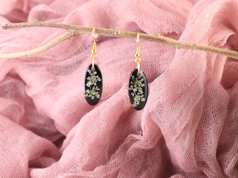 May include: A pair of black oval earrings with gold hooks. Each earring features a delicate white flower pressed inside the resin.