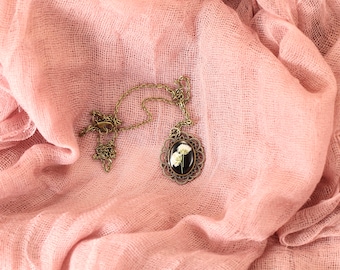 Pode incluir: Um delicado colar de corrente dourada com um pingente redondo com um design de flores prensadas. O pingente tem um fundo preto e flores brancas.