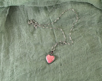 May include: A silver necklace with a heart-shaped pendant. The pendant has a pink border and a light pink, textured center. The necklace chain is delicate and lies on a sage green fabric.