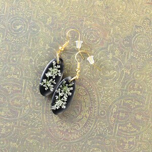 May include: A pair of black oval earrings with gold hooks. Each earring features a delicate white flower pressed inside the resin.