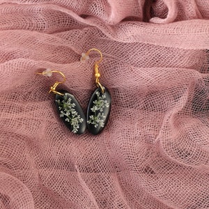 May include: A pair of black resin earrings with gold hooks. The earrings are oval shaped and feature delicate white flowers pressed into the resin.