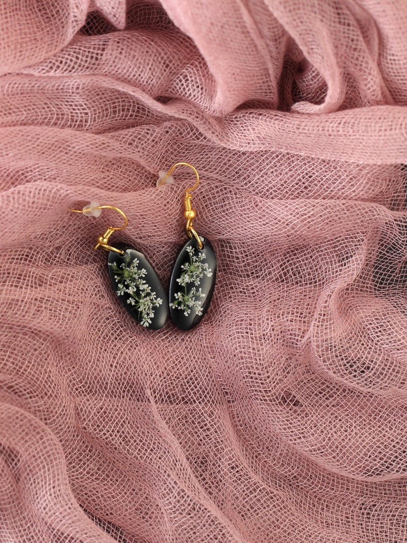 May include: A pair of black resin earrings with gold hooks. The earrings are oval shaped and feature delicate white flowers pressed into the resin.