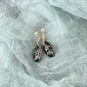 May include: A pair of black resin earrings with delicate white flowers pressed inside. The earrings are oval shaped and have gold hooks.