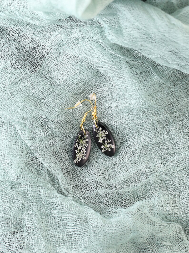 May include: A pair of black resin earrings with delicate white flowers pressed inside. The earrings are oval shaped and have gold hooks.