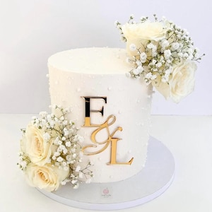 May include: A white wedding cake decorated with white flowers and gold letters spelling "E & L".