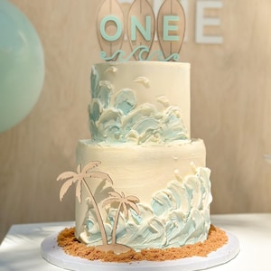 Op de afbeelding: Een tweelaagse witte taart versierd met blauwe en witte frosting om op oceaan golven te lijken. De taart is bedekt met een houten surfboard met het woord "ONE" erop. Er zijn ook twee houten palmbomen op de onderste laag van de taart.