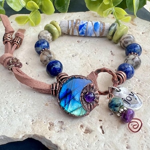 Könnte beinhalten: Handgefertigtes Armband mit einem blauen und violetten Edelsteinanhänger und Kupferakzenten. Das Armband enthält blaue, grüne und graue Perlen, einen kleinen "joy" Anhänger und eine Wildleder-Kordel.