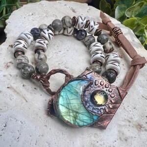 May include: A copper necklace with a large, iridescent labradorite stone pendant surrounded by smaller stones. The necklace is strung with white and gray beads and a brown leather cord.