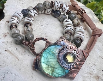 May include: A copper necklace with a large, iridescent labradorite stone pendant surrounded by smaller stones. The necklace is strung with white and gray beads and a brown leather cord.