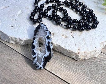 May include: A black onyx beaded necklace with a large, white and black agate pendant. The pendant has a natural, raw edge and a hole drilled through the top for stringing.