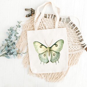 Cream tote bag with a large green Luna moth illustration displayed in a styled flat lay with eucalyptus, macramé, and a rustic stick on a white wood background.