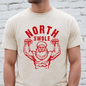 May include: A cream-colored t-shirt with a red graphic of a muscular Santa Claus with the text "NORTH SWOLE" above him.