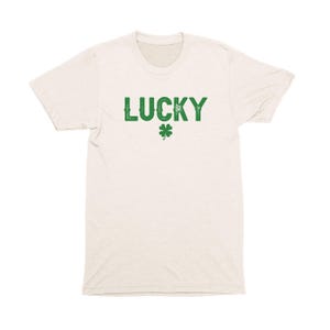 Op de afbeelding: Een licht beige t-shirt met het woord "LUCKY" in het groen bedrukt, met daaronder een groen klavertje vier.