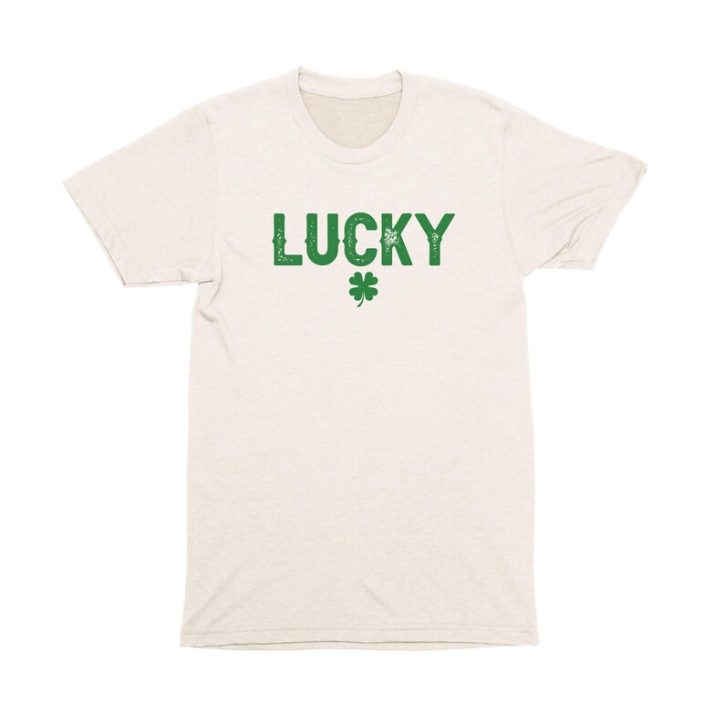 Op de afbeelding: Een licht beige t-shirt met het woord "LUCKY" in het groen bedrukt, met daaronder een groen klavertje vier.
