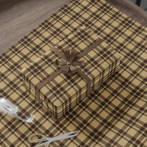 May include: A brown and tan plaid gift wrap with a brown ribbon and bow on top of a gift box. The gift box is also wrapped in brown and tan plaid paper.