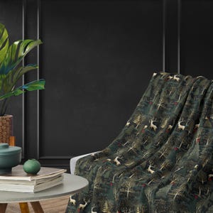 May include: A green throw blanket with a repeating pattern of trees, deer, and red berries. The blanket is draped over a chair in a room with a dark grey wall.