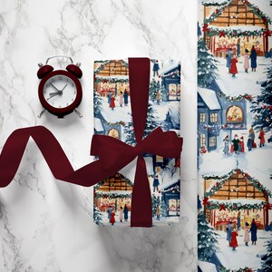 Puede incluir: Una caja de regalo roja y blanca con un diseño festivo de pueblo navideño. La caja está atada con una cinta color burdeos y un reloj despertador rojo está en el fondo.
