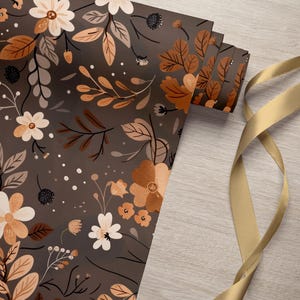 May include: A roll of brown wrapping paper with a floral pattern featuring white and orange flowers and leaves. A gold ribbon is lying on a wooden surface.