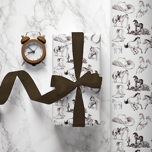 May include: A white gift box wrapped with brown ribbon and a cowboy-themed pattern of horses, boots, hats, and horseshoes. The box is on a white marble surface with a brown alarm clock beside it.