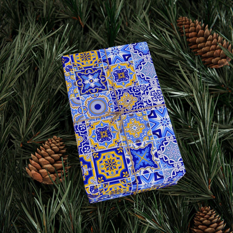May include: A blue and yellow gift box with a geometric pattern, wrapped with twine and tied in a bow. The box is sitting on a bed of green pine needles.