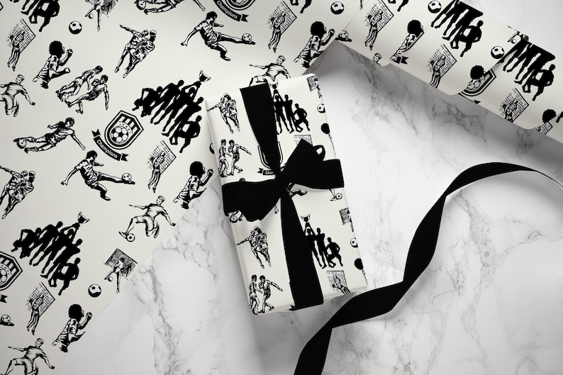 May include: Black and white wrapping paper with a repeating pattern of soccer players, soccer balls, and trophies. The paper is wrapped around a gift box with a black ribbon.