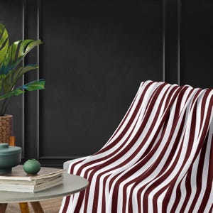 May include: A white and maroon striped throw blanket draped over a chair. The blanket is made of soft, plush fabric and has a classic striped pattern.
