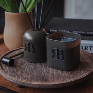May include: A dark grey aromatherapy diffuser and a dark grey scented candle with the text "Relics & Rituals" and the initials "RB" on both. The diffuser is on a wooden tray with a black metal lid.
