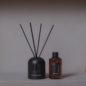 May include: A matte black aromatherapy diffuser with four black reed sticks and a matching amber glass bottle with a black cap. The black diffuser features the letters RB and the text AROMATHERAPY DIFFUSER. The amber bottle has a black label with Scent Bloom.