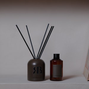 May include: A brown ceramic diffuser bottle with black reeds and a black label with the text "RB" and "ROOM & BOUDOIR" on a shelf with a brown glass bottle with a black cap and a stack of books in the background.