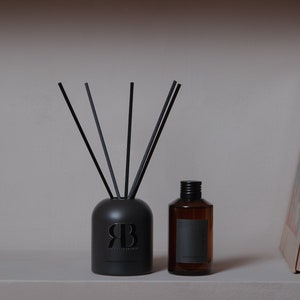Peut inclure: Diffuseur noir avec des tiges noires et une étiquette noire avec le texte "RETROROMANTIQUE" et la lettre "R" dans une police stylisée. Un petit flacon en verre brun avec un bouchon noir est à côté du diffuseur.